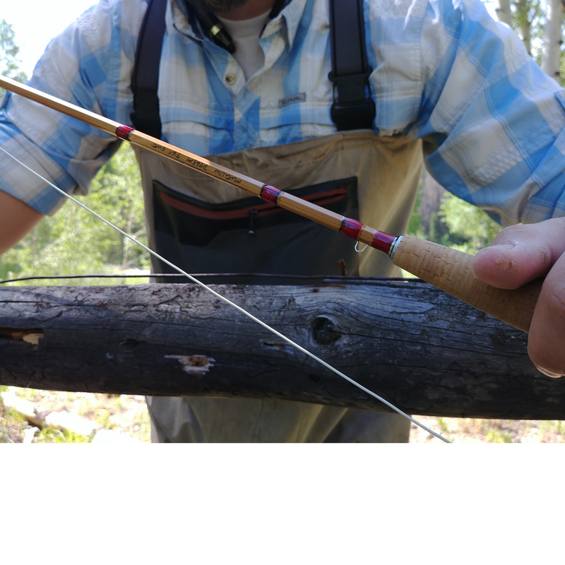 close-up of a bamboo cane fly rod with red thread wraps and cork grip, held over a log with line out